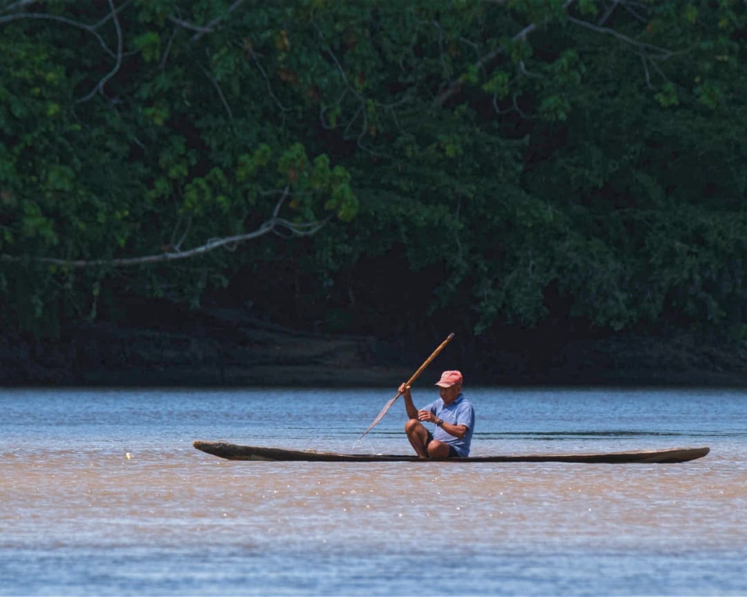 A man paddles a traditional wooden canoe on the deep blue-gray river backed by dense jungle foliage