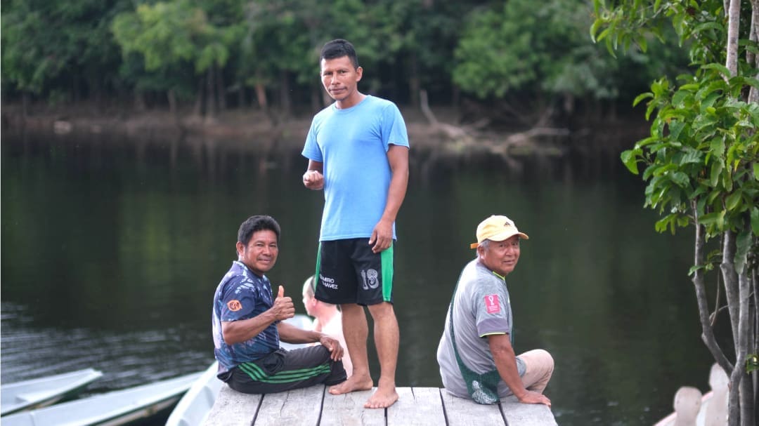 Three men on a dock, two are sitting with their legs hanging over the edge; a third stands in between; two men are holding their hands in a thumbs up position, all are smiling