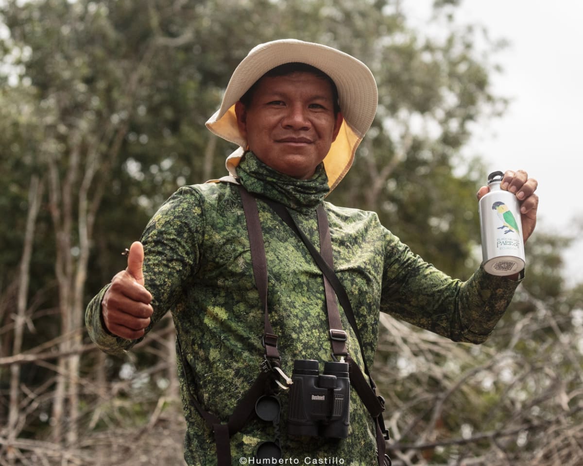 Kichwa man wearing birdwatching gear holds a water bottle in his left hand and gives a thumbs up with his right
