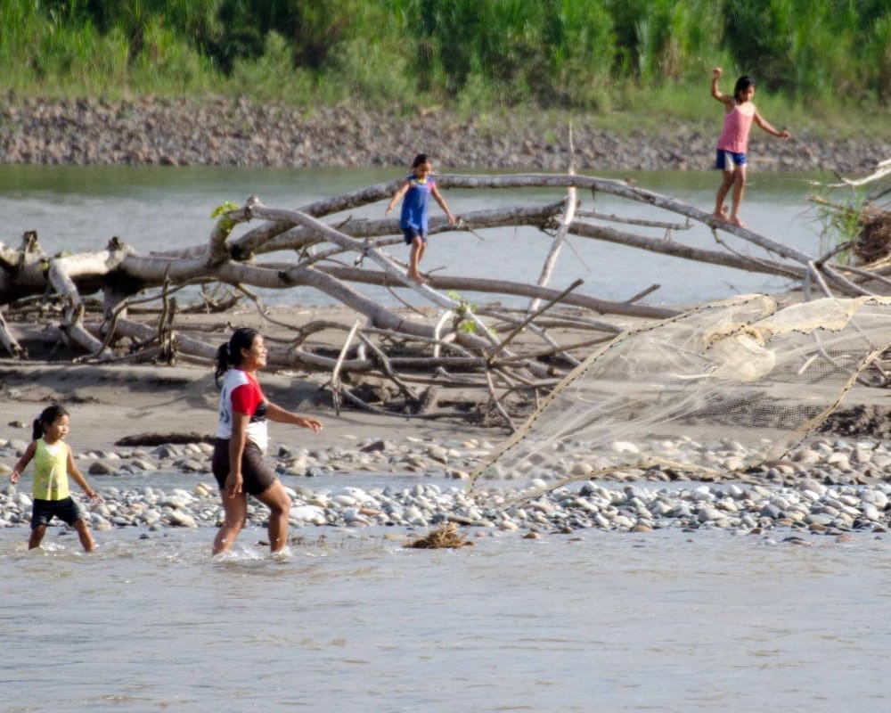 A woman tosses a weighted fishing net into the river while her children play nearby